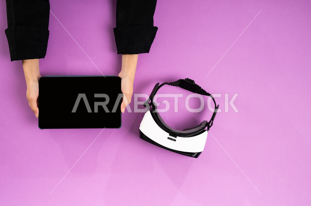 الاستمتاع في العاب الفيديو، عرض شاشة سوداء فارغة، بورتريه علوي عامودي ليدين إمرأة عربية خليجية سعودية تعمل على التابليت وبجانبها نظارة الفي آر وقبضة التحكم، استخدام الأجهزة التقنية الحديثة, محاكاة العالم الافتراضي، التطور التكنولوجي في المملكة، خلفية وردية