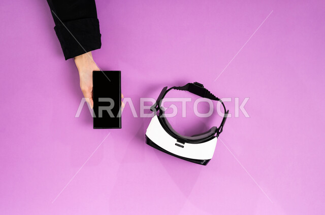 Technological development in the Kingdom, enjoying video games, displaying a blank black screen, vertical overhead portrait of the hands of a Saudi Arabian Gulf woman working on a mobile phone and next to her are VR glasses and a joystick, using modern technical devices, simulating the virtual world, pink background