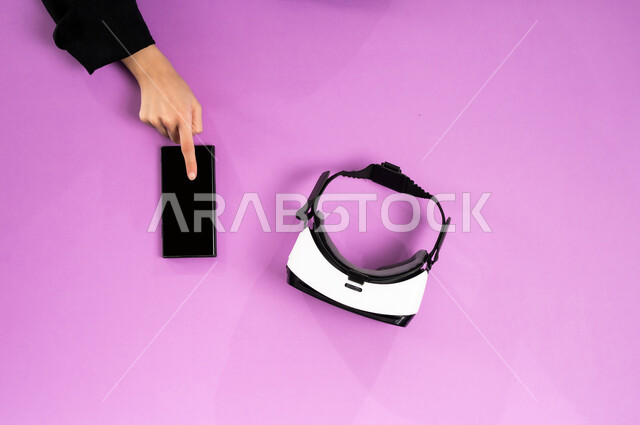 Virtual world simulation, technological development in the Kingdom, enjoying video games, displaying a blank black screen, vertical top portrait of the hands of a Saudi Arabian Gulf woman working on a mobile phone and next to her are VR glasses and a joystick, using modern technical devices, pink background