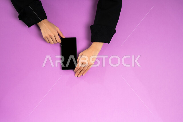 استخدام التقنيات الحديثة والمتطورة بالعمل، البيع والشراء عبر الانترنت، الاستفادة من تطبيقات التسوق أونلاين، عرض شاشة سوداء فارغة، إنجاز المهام المطلوبة عن بعد، بورتريه علوي عامودي ليدين إمرأة عربية خليجية سعودية تعمل على جهاز الهاتف المحمول، خلفية وردية