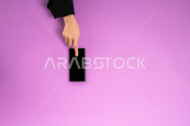 إنجاز المهام المطلوبة عن بعد، بورتريه علوي عامودي ليدين إمرأة عربية خليجية سعودية تعمل على جهاز الهاتف المحمول، استخدام التقنيات الحديثة والمتطورة بالعمل، البيع والشراء عبر الانترنت، الاستفادة من تطبيقات التسوق أونلاين، عرض شاشة سوداء فارغة، خلفية وردية