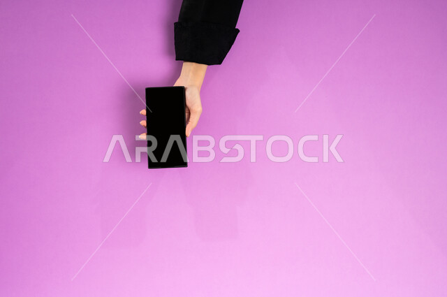 الاستفادة من تطبيقات التسوق أونلاين، عرض شاشة سوداء فارغة، إنجاز المهام المطلوبة عن بعد، بورتريه علوي عامودي ليدين إمرأة عربية خليجية سعودية تعمل على جهاز الهاتف المحمول، استخدام التقنيات الحديثة والمتطورة بالعمل، البيع والشراء عبر الانترنت، خلفية وردية