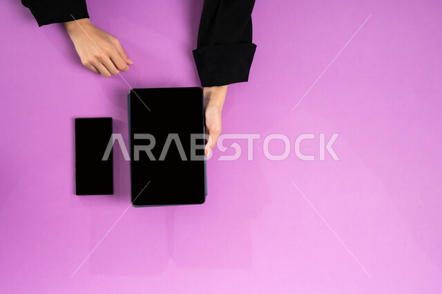 مهن ووظائف مكتبية سعودية، دمج العمل مع التقنية والتكنولوجيا الحديثة، مفهوم ادارة الاعمال وتحقيق الأهداف أونلاين، إنجاز المهام المطلوبة عن بعد، بورتريه علوي عامودي ليدين إمرأة عربية خليجية سعودية تعمل على التابليت والجوال، عرض شاشة سوداء فارغة، خلفية وردية