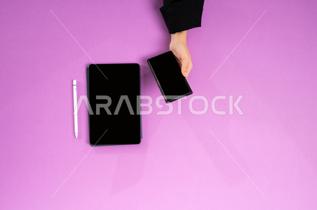 مفهوم ادارة الاعمال وتحقيق الأهداف أونلاين، إنجاز المهام المطلوبة عن بعد، بورتريه علوي عامودي ليدين إمرأة عربية خليجية سعودية تعمل على التابليت والجوال، عرض شاشة سوداء فارغة، مهن ووظائف مكتبية سعودية، دمج العمل مع التقنية والتكنولوجيا الحديثة، خلفية وردية