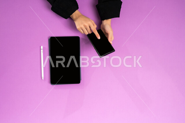 Integrating work with technology and modern technology, the concept of business management and achieving goals online, accomplishing the required tasks remotely, vertical upper portrait of the hands of a Saudi Arabian Gulf woman working on a tablet and mobile, displaying a blank black screen, Saudi office jobs and professions, pink background