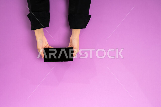 استخدام التقنيات الحديثة والمتطورة بالعمل، البيع والشراء عبر الانترنت، الاستفادة من تطبيقات التسوق أونلاين، عرض شاشة سوداء فارغة، إنجاز المهام المطلوبة عن بعد، بورتريه علوي عامودي ليدين إمرأة عربية خليجية سعودية تعمل على جهاز الهاتف المحمول، خلفية وردية