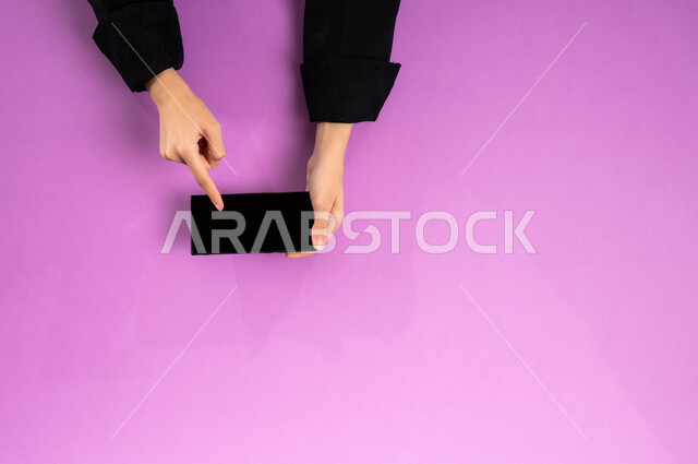 إنجاز المهام المطلوبة عن بعد، بورتريه علوي عامودي ليدين إمرأة عربية خليجية سعودية تعمل على جهاز الهاتف المحمول، استخدام التقنيات الحديثة والمتطورة بالعمل، البيع والشراء عبر الانترنت، الاستفادة من تطبيقات التسوق أونلاين، عرض شاشة سوداء فارغة، خلفية وردية