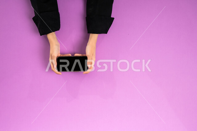 Displaying a blank black screen, accomplishing the required tasks remotely, a vertical upper portrait of the hands of a Saudi Arabian Gulf woman working on a mobile phone, using modern and advanced technologies at work, buying and selling online, benefiting from online shopping applications, pink background
