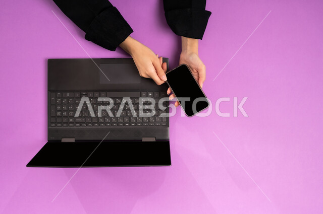 عرض شاشة سوداء فارغة، مهن ووظائف مكتبية سعودية، دمج العمل مع التقنية والتكنولوجيا الحديثة، مفهوم ادارة الاعمال وتحقيق الأهداف أونلاين، إنجاز المهام المطلوبة عن بعد، بورتريه علوي عامودي ليدين إمرأة عربية خليجية سعودية تعمل على  اللابتوب والجوال، خلفية وردية