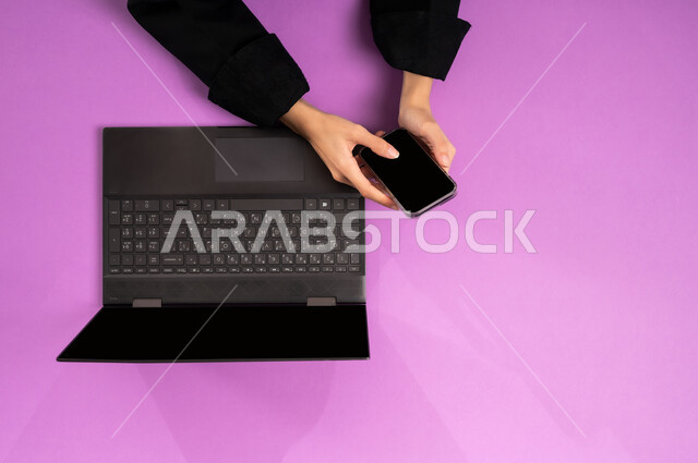 إنجاز المهام المطلوبة عن بعد، بورتريه علوي عامودي ليدين إمرأة عربية خليجية سعودية تعمل على  اللابتوب والجوال، عرض شاشة سوداء فارغة، مهن ووظائف مكتبية سعودية، دمج العمل مع التقنية والتكنولوجيا الحديثة، مفهوم ادارة الاعمال وتحقيق الأهداف أونلاين، خلفية وردية