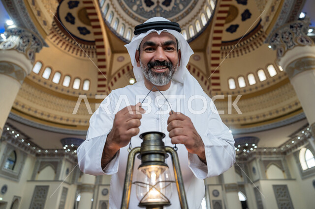 الاحتفال بقدوم شهر رمضان المبارك, العبادة والتقرب إلى الله, أداء الفرائض الدينية, التصاميم والديكورات الداخلية للمساجد في الإمارات، رجل عربي خليجي إماراتي يرتدي الكندورة والغترة البيضاء يقف في المسجد يمسك بيده فانوس وينظر إلى الكاميرا بإيماءات السعادة والسرور