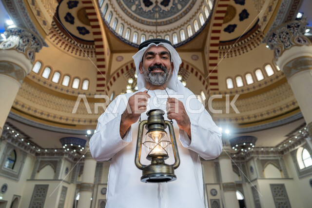 Worship and getting closer to God, performing religious duties, interior designs and decorations of mosques in the Emirates, celebrating the arrival of the holy month of Ramadan, an Emirati Gulf Arab man wearing a white kandura and ghutra stands in the mosque holding a lantern in his hand and looking at the camera with gestures of happiness and pleasure