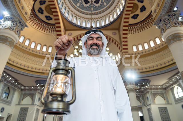 التصاميم والديكورات الداخلية للمساجد في الإمارات، أداء الفرائض الدينية, العبادة والتقرب إلى الله, الاحتفال بقدوم شهر رمضان المبارك، رجل عربي خليجي إماراتي يرتدي الكندورة والغترة البيضاء يقف في المسجد يمسك بيده فانوس وينظر إلى الكاميرا بإيماءات السعادة والسرور