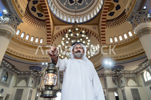 الاحتفال بقدوم شهر رمضان المبارك، التصاميم والديكورات الداخلية للمساجد في الإمارات، أداء الفرائض الدينية, العبادة والتقرب إلى الله، رجل عربي خليجي إماراتي يرتدي الكندورة والغترة البيضاء يقف في المسجد يمسك بيده فانوس وينظر إلى الكاميرا بإيماءات السعادة والسرور