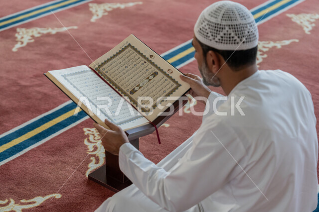 مفهوم العبادة والطاعة والتقرب الى الله, التمعن بسور القرآن الكريم, تدبر الآيات القرآنية والتفكر بها، الالتزام بالورد اليومي, صورة مقربة من الخلف لرجل عربي خليجي إماراتي يرتدي الكندورة والقبعة البيضاء يجلس في المسجد ويقرأ من المصحف الشريف, التعبد في بيوت الرحمن