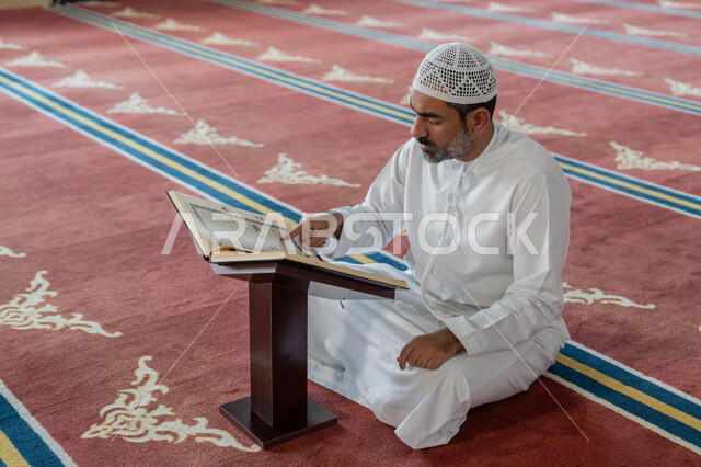 التعبد في بيوت الرحمن, التمعن بسور القرآن الكريم، مفهوم العبادة والطاعة والتقرب الى الله, تدبر الآيات القرآنية والتفكر بها، الالتزام بالورد اليومي, رجل عربي خليجي إماراتي يرتدي الكندورة والقبعة البيضاء يجلس في المسجد ويقرأ من المصحف الشريف