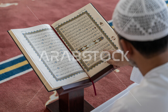 التمعن بسور القرآن الكريم، مفهوم العبادة والطاعة والتقرب الى الله, تدبر الآيات القرآنية والتفكر بها، الالتزام بالورد اليومي, صورة مقربة من الخلف رجل عربي خليجي إماراتي يرتدي الكندورة والقبعة البيضاء يجلس في المسجد ويقرأ من المصحف الشريف, التعبد في بيوت الرحمن