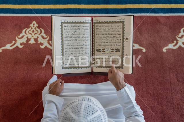 الالتزام بالورد اليومي, التمعن بسور القرآن الكريم، مفهوم العبادة والطاعة والتقرب الى الله, تدبر الآيات القرآنية والتفكر بها, صورة من الأعلى لرجل عربي خليجي إماراتي يرتدي الكندورة والقبعة البيضاء يجلس في المسجد ويقرأ من المصحف الشريف, التعبد في بيوت الرحمن