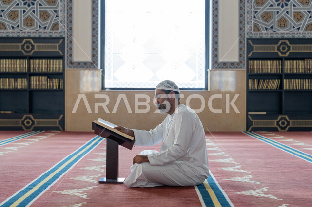 The concept of worship, obedience and drawing closer to God, contemplating the chapters of the Holy Quran, pondering and reflecting on the verses of the Quran, committing to the daily rosary, a side view of an Arab Gulf Emirati man wearing a kandura and a white hat sitting in the mosque and reading from the Holy Quran, worship in the houses of the Most Merciful