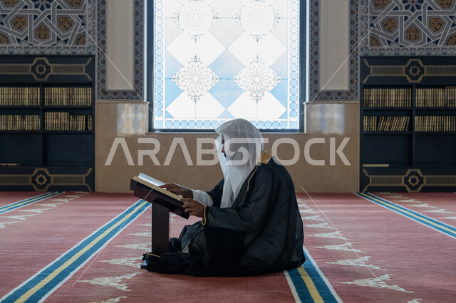 التمعن بسور القرآن الكريم، مفهوم العبادة والتقرب الى الله, تدبر الآيات القرآنية والتفكر بها، الالتزام بالورد اليومي, صورة من الجانب لرجل عربي خليجي إماراتي يرتدي البشت والكندورة والغترة البيضاء يجلس في المسجد ويقرأ من المصحف الشريف, التعبد في بيوت الرحمن