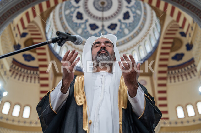 مفهوم الخشوع والتضرع, طلب الرحمة والمغفرة, إمامة المسلمين في المسجد، العبادة والتقرب إلى الله، تعزيز الروحانية الجماعية, إمام عربي خليجي إماراتي يرتدي البشت والكندورة والغترة البيضاء يقوم بأداء فريضة الصلاة ويرفع يديه بتعابير الدعاء
