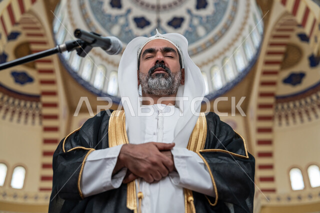 تعزيز الروحانية الجماعية, أداء الفرائض الدينية الإسلامية, مفهوم أركان الإسلام, العبادة والتقرب إلى الله, إمامة المسلمين في المسجد, صورة مقربة لإمام عربي خليجي إماراتي يرتدي البشت والكندورة والغترة البيضاء يقوم بأداء فريضة الصلاة ويضع كفه اليمنى فوق اليسرى