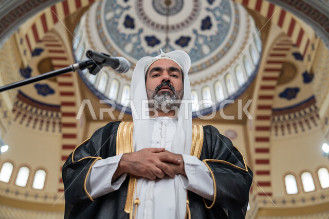 أداء الفرائض الدينية الإسلامية, مفهوم أركان الإسلام, تعزيز الروحانية الجماعية, العبادة والتقرب إلى الله, إمامة المسلمين في المسجد, إمام عربي خليجي إماراتي يرتدي البشت والكندورة والغترة البيضاء يقوم بأداء فريضة الصلاة ويضع كفه اليمنى فوق اليسرى