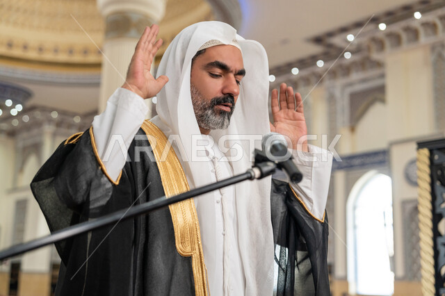 العبادة والتقرب إلى الله, إمامة المسلمين في المسجد، تعزيز الروحانية الجماعية, مفهوم أركان الإسلام، أداء الفرائض الدينية الإسلامية, صورة مقربة لإمام عربي خليجي إماراتي يرتدي البشت والكندورة والغترة البيضاء يقوم بأداء فريضة الصلاة ويرفع يديه لتكبيرة الإحرام