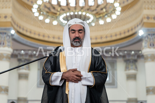 أداء الفرائض الدينية الإسلامية, مفهوم أركان الإسلام, تعزيز الروحانية الجماعية, العبادة والتقرب إلى الله, إمامة المسلمين في المسجد, إمام عربي خليجي إماراتي يرتدي البشت والكندورة والغترة البيضاء يقوم بأداء فريضة الصلاة ويضع كفه اليمنى فوق اليسرى