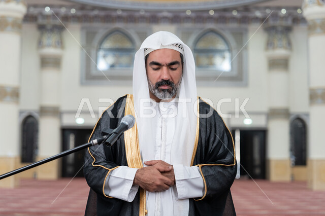 مفهوم أركان الإسلام، أداء الفرائض الدينية الإسلامية, تعزيز الروحانية الجماعية, العبادة والتقرب إلى الله, إمامة المسلمين في المسجد, صورة مقربة لإمام عربي خليجي إماراتي يرتدي البشت والكندورة والغترة البيضاء يقوم بأداء فريضة الصلاة ويضع كفه اليمنى فوق اليسرى
