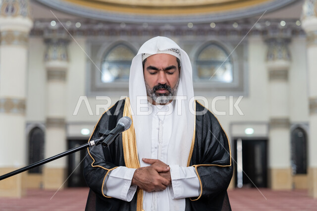 تعزيز الروحانية الجماعية, مفهوم أركان الإسلام، أداء الفرائض الدينية الإسلامية, العبادة والتقرب إلى الله, إمامة المسلمين في المسجد, صورة مقربة لإمام عربي خليجي إماراتي يرتدي البشت والكندورة والغترة البيضاء يقوم بأداء فريضة الصلاة ويضع كفه اليمنى فوق اليسرى