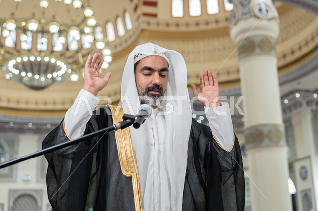 إمامة المسلمين في المسجد، العبادة والتقرب إلى الله، تعزيز الروحانية الجماعية, مفهوم أركان الإسلام، أداء الفرائض الدينية الإسلامية, صورة مقربة لإمام عربي خليجي إماراتي يرتدي البشت والكندورة والغترة البيضاء يقوم بأداء فريضة الصلاة ويرفع يديه لتكبيرة الإحرام
