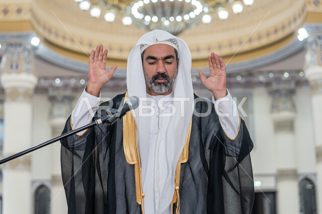 أداء فرائض الدين الإسلامي، الركن الثاني من أركان الإسلام, الشعور بالخشوع والطمأنينة, إمامة المسلمين في المسجد, إقامة الصلاة على وقتها، صورة مقربة لإمام عربي خليجي إماراتي يرتدي البشت والكندورة والغترة البيضاء يقوم بأداء فريضة الصلاة ويرفع يديه لتكبيرة الإحرام