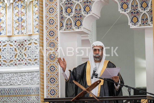 مفهوم العبادة والتقرب إلى الله تعالى, حث المسلمين لأداء الفرائض, توجيه الناس نحو الحق والهداية, دور أئمة المساجد في نشر الدين، إمام مسجد عربي خليجي إماراتي يرتدي البشت والكندورة والغترة البيضاء يقف على المنبر يمسك بيده أوراق ويلقي خطبة يوم الجمعة