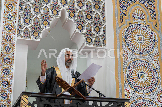 حث المسلمين لأداء الفرائض, توجيه الناس نحو الحق والهداية, مفهوم العبادة والتقرب إلى الله تعالى, دور أئمة المساجد في نشر الدين، إمام مسجد عربي خليجي إماراتي يرتدي البشت والكندورة والغترة البيضاء يقف على المنبر يمسك بيده أوراق ويلقي خطبة يوم الجمعة