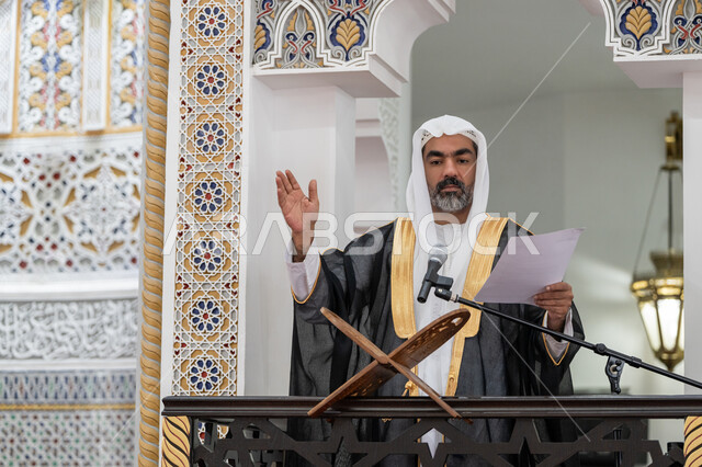 مفهوم العبادة والتقرب إلى الله تعالى, توجيه الناس نحو الحق والهداية, حث المسلمين لأداء الفرائض, دور أئمة المساجد في نشر الدين، صورة مقربة لإمام مسجد عربي خليجي إماراتي يرتدي البشت والكندورة والغترة البيضاء يقف على المنبر يمسك بيده أوراق ويلقي خطبة يوم الجمعة