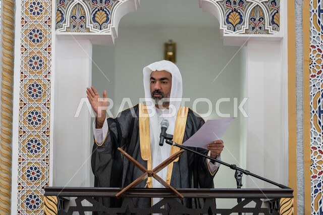 دور أئمة المساجد في نشر الدين, مفهوم العبادة والتقرب إلى الله تعالى, توجيه الناس نحو الحق والهداية, حث المسلمين لأداء الفرائض، صورة مقربة لإمام مسجد عربي خليجي إماراتي يرتدي البشت والكندورة والغترة البيضاء يقف على المنبر يمسك بيده أوراق ويلقي خطبة يوم الجمعة