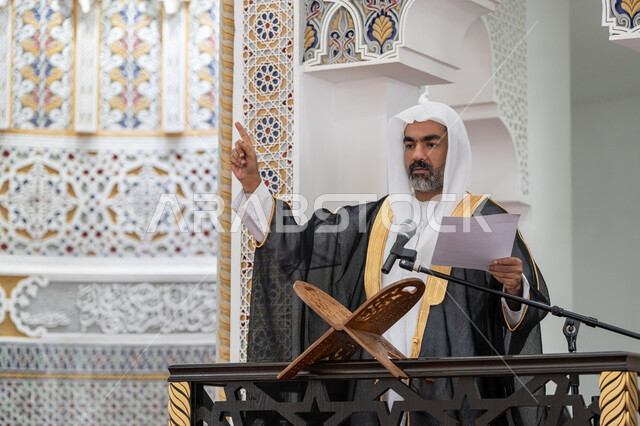 حث المسلمين لأداء الفرائض, توجيه الناس نحو الحق والهداية, مفهوم العبادة والتقرب إلى الله تعالى, دور أئمة المساجد في نشر الدين، صورة مقربة لإمام مسجد عربي خليجي إماراتي يرتدي البشت والكندورة والغترة البيضاء يقف على المنبر يمسك بيده أوراق ويلقي خطبة يوم الجمعة