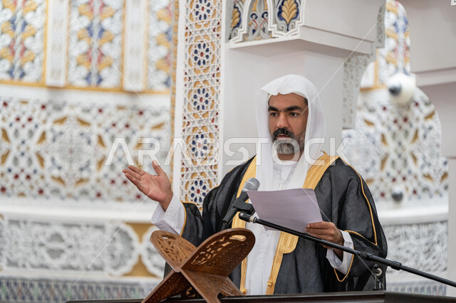 Guiding people towards truth and guidance, urging Muslims to perform the obligatory duties, the concept of worship and drawing closer to God Almighty, the role of mosque imams in spreading religion, a close-up photo of an Emirati Gulf Arab mosque imam wearing a bisht, kandura and white ghutra standing on the pulpit holding papers in his hand and delivering a Friday sermon