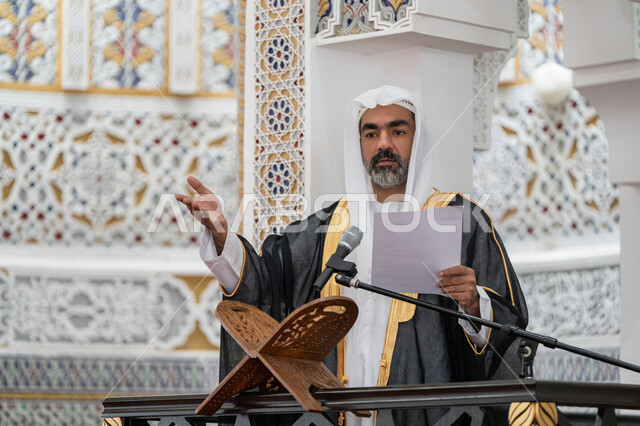 مفهوم العبادة والتقرب إلى الله تعالى, توجيه الناس نحو الحق والهداية, حث المسلمين لأداء الفرائض, دور أئمة المساجد في نشر الدين، صورة مقربة لإمام مسجد عربي خليجي إماراتي يرتدي البشت والكندورة والغترة البيضاء يقف على المنبر يمسك بيده أوراق ويلقي خطبة يوم الجمعة