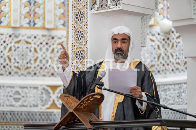 دور أئمة المساجد في نشر الدين, مفهوم العبادة والتقرب إلى الله تعالى, توجيه الناس نحو الحق والهداية, حث المسلمين لأداء الفرائض، صورة مقربة لإمام مسجد عربي خليجي إماراتي يرتدي البشت والكندورة والغترة البيضاء يقف على المنبر يمسك بيده أوراق ويلقي خطبة يوم الجمعة