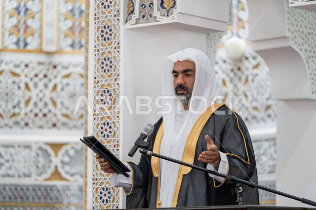العبادة والتقرب إلى الله, الحث على أداء الفرائض, توجيه الناس لطريق الحق والهداية, إلقاء خطاب و دروس توجيهية على المسلمين, صورة مقربة لإمام مسجد عربي خليجي إماراتي يرتدي البشت والكندورة والغترة البيضاء يقف على المنبر ويلقي خطبة يوم الجمعة من الجهاز اللوحي