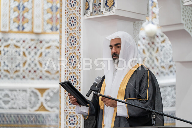 الحث على أداء العبادات والفرائض الدينية, توجيه الناس لطريق الحق والهداية, إلقاء خطاب و دروس توجيهية على المسلمين, صورة مقربة لإمام مسجد عربي خليجي إماراتي يرتدي البشت والكندورة والغترة البيضاء يقف على المنبر ويلقي خطبة يوم الجمعة من الجهاز اللوحي