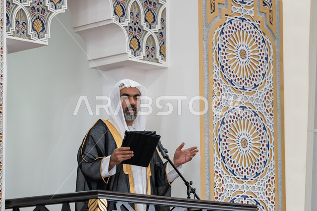 إلقاء خطاب و دروس توجيهية على المسلمين, توجيه الناس لطريق الحق والهداية, الحث على أداء العبادات والفرائض الدينية, إمام مسجد عربي خليجي إماراتي يرتدي البشت والكندورة والغترة البيضاء يقف على المنبر ويلقي خطبة يوم الجمعة من الجهاز اللوحي