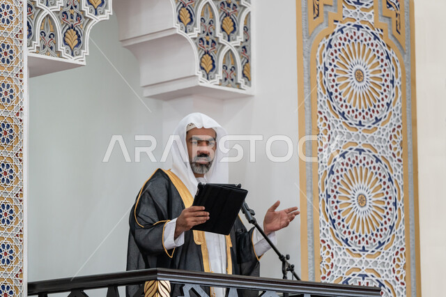 العبادة والتقرب إلى الله, الحث على أداء العبادات والفرائض الدينية, توجيه الناس لطريق الحق والهداية, إلقاء خطاب و دروس توجيهية على المسلمين, إمام مسجد عربي خليجي إماراتي يرتدي البشت والكندورة والغترة البيضاء يقف على المنبر ويلقي خطبة يوم الجمعة من الجهاز اللوحي