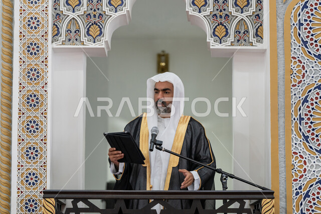 الحث على أداء العبادات والفرائض الدينية, توجيه الناس لطريق الحق والهداية, إلقاء خطاب و دروس توجيهية على المسلمين, إمام مسجد عربي خليجي إماراتي يرتدي البشت والكندورة والغترة البيضاء يقف على المنبر ويلقي خطبة يوم الجمعة من الجهاز اللوحي