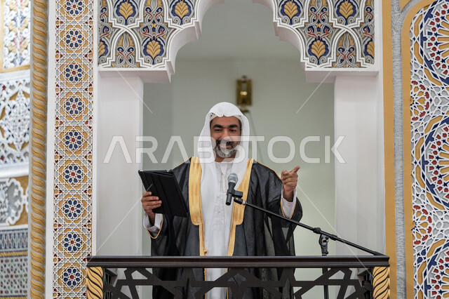 توجيه الناس لطريق الحق والهداية, الحث على أداء العبادات والفرائض الدينية, إلقاء خطاب و دروس توجيهية على المسلمين, إمام مسجد عربي خليجي إماراتي يرتدي البشت والكندورة والغترة البيضاء يقف على المنبر ويلقي خطبة يوم الجمعة من الجهاز اللوحي