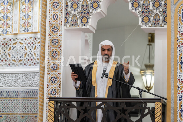 إلقاء خطاب و دروس توجيهية على المسلمين, توجيه الناس لطريق الحق والهداية, الحث على أداء العبادات والفرائض الدينية, إمام مسجد عربي خليجي إماراتي يرتدي البشت والكندورة والغترة البيضاء يقف على المنبر ويلقي خطبة يوم الجمعة من الجهاز اللوحي