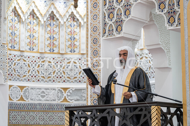 Guiding people to the path of truth and guidance, urging them to perform religious duties and rituals, giving a speech and guidance lessons to Muslims, an imam of an Arab Gulf Emirati mosque wearing a bisht, kandura and white ghutra stands on the pulpit and delivers the Friday sermon from a tablet