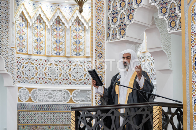 إلقاء خطاب و دروس توجيهية على المسلمين, توجيه الناس لطريق الحق والهداية, الحث على أداء العبادات والفرائض الدينية, إمام مسجد عربي خليجي إماراتي يرتدي البشت والكندورة والغترة البيضاء يقف على المنبر ويلقي خطبة يوم الجمعة من الجهاز اللوحي